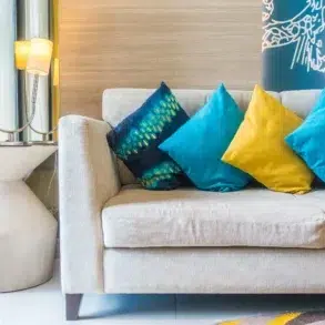 Beige sofa with blue and yellow decorative pillows, next to a white side table with a lamp and yellow curtains.