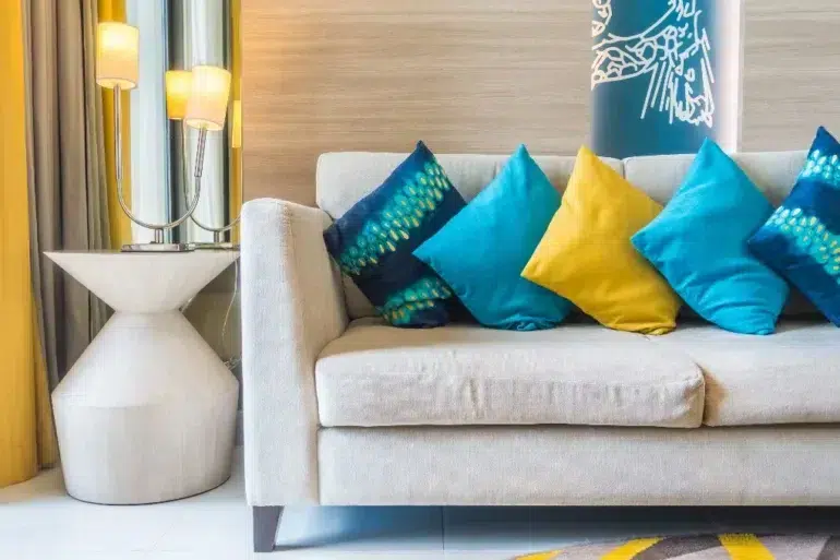 Beige sofa with blue and yellow decorative pillows, next to a white side table with a lamp and yellow curtains.