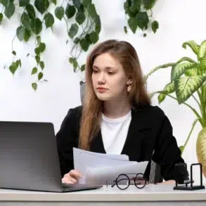 A person sits at a desk with a laptop, holding documents, surrounded by greenery and an office globe.