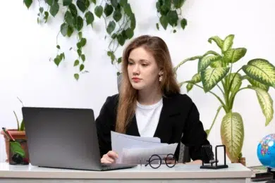 A person sits at a desk with a laptop, holding documents, surrounded by greenery and an office globe.
