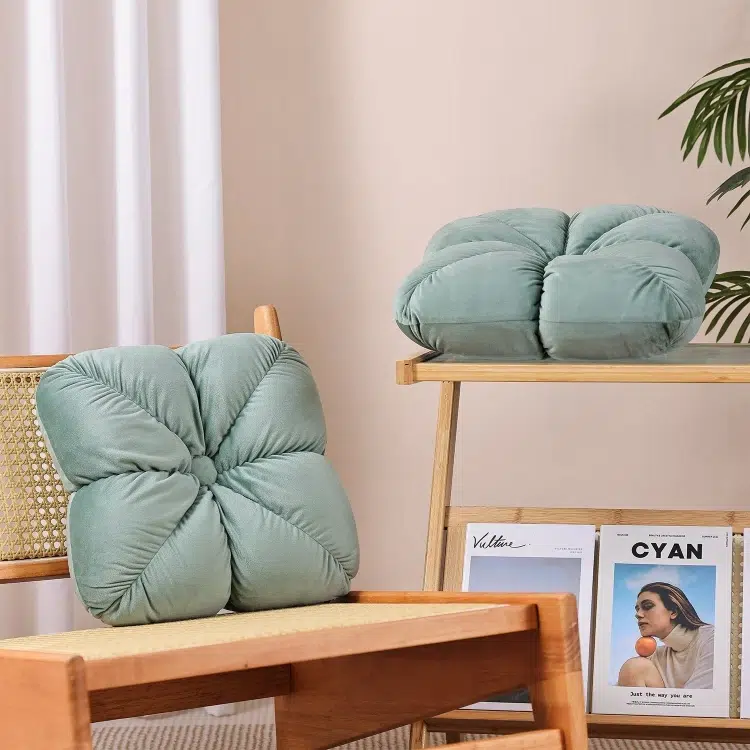 A cozy scene with two tufted green pillows, one on a woven chair and the other on a wooden shelf. Magazines are displayed below, with a plant nearby.