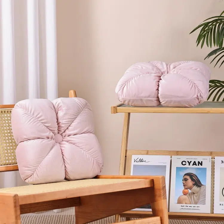 Cozy room with two blush pink flower-shaped cushions; one on a chair, another on a wooden shelf with books and a potted plant nearby. Soft and inviting.