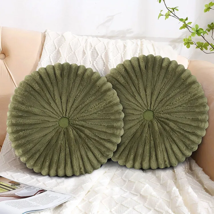 Two round, plush green pillows with a pleated design rest on a white textured blanket atop a beige couch. A magazine and green plant add cozy accents.