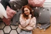 A woman with red hair smiles gently, lying on a patterned rug surrounded by soft, pink and gray pillows in a cozy setting. Warm, relaxed ambiance.