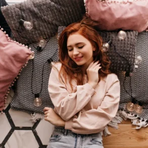 A woman with red hair smiles gently, lying on a patterned rug surrounded by soft, pink and gray pillows in a cozy setting. Warm, relaxed ambiance.
