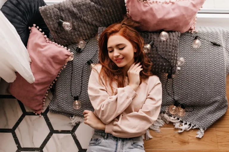 A woman with red hair smiles gently, lying on a patterned rug surrounded by soft, pink and gray pillows in a cozy setting. Warm, relaxed ambiance.