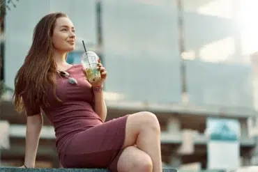 A woman in a mauve dress sits on a ledge, sipping a drink with a straw. She appears relaxed and content, with a cityscape blurred in the background.