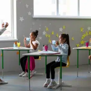 A teacher stands in front of students at desks, engaged in a lesson while children hold materials and participate actively in class.