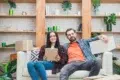 A smiling couple sits on a couch holding boxes, surrounded by houseplants and shelves, suggesting a cheerful moving or unpacking scene.