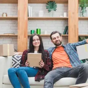 A smiling couple sits on a couch holding boxes, surrounded by houseplants and shelves, suggesting a cheerful moving or unpacking scene.