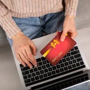 Person holding a red gift card with a yellow ribbon over a laptop keyboard, wearing a beige sweater and blue jeans.