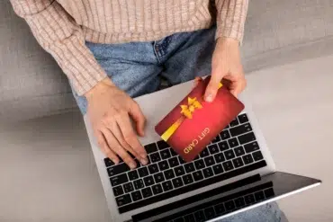 Person holding a red gift card with a yellow ribbon over a laptop keyboard, wearing a beige sweater and blue jeans.
