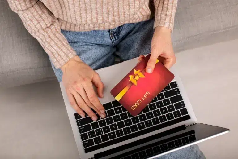 Person holding a red gift card with a yellow ribbon over a laptop keyboard, wearing a beige sweater and blue jeans.