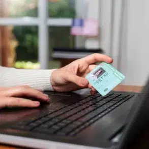 Person holding a U.S. driver's license near a laptop keyboard, suggesting online identity verification or digital access.