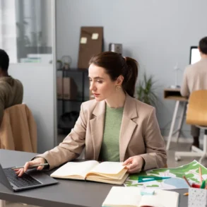 A woman seated at a desk works on a laptop, with notebooks and a colorful map spread out, while two others are focused at their computers.