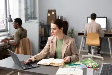 A woman seated at a desk works on a laptop, with notebooks and a colorful map spread out, while two others are focused at their computers.