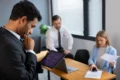 Three business professionals in a modern office reviewing charts and data on a tablet, laptop, and printed papers during a meeting.