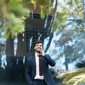 A smiling man in a navy suit talks on a phone outdoors. He stands near a reflective glass wall with trees and leaves visible, conveying a relaxed, professional mood.