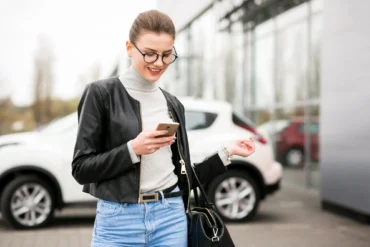 A person in a black leather jacket and gray sweater stands outside, looking at their phone near parked cars.