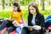 A group of young people sitting on a red checkered blanket outdoors in a grassy area with trees, studying and using a tablet.