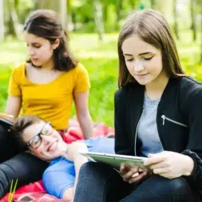 A group of young people sitting on a red checkered blanket outdoors in a grassy area with trees, studying and using a tablet.