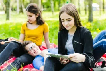 A group of young people sitting on a red checkered blanket outdoors in a grassy area with trees, studying and using a tablet.