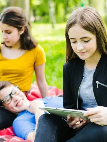 A group of young people sitting on a red checkered blanket outdoors in a grassy area with trees, studying and using a tablet.