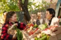 A woman in a red plaid shirt holds fresh carrots while conversing with a vendor at an outdoor market stand full of colorful produce, under sunlit trees.