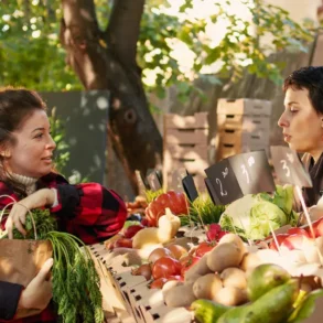 A woman in a red plaid shirt holds fresh carrots while conversing with a vendor at an outdoor market stand full of colorful produce, under sunlit trees.