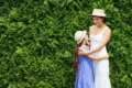 A woman and a young girl in straw hats and dresses happily embrace in front of a lush green hedge, conveying warmth and affection.