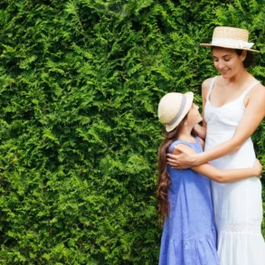 A woman and a young girl in straw hats and dresses happily embrace in front of a lush green hedge, conveying warmth and affection.