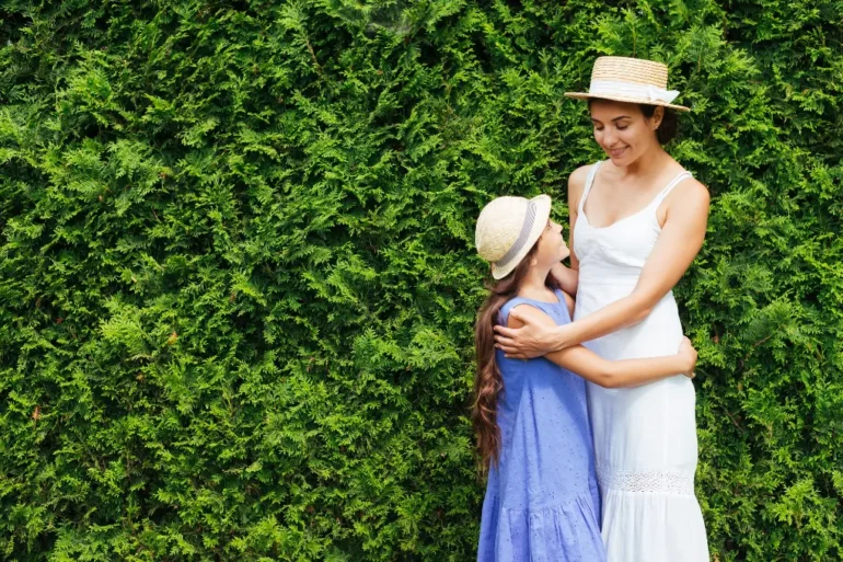 A woman and a young girl in straw hats and dresses happily embrace in front of a lush green hedge, conveying warmth and affection.