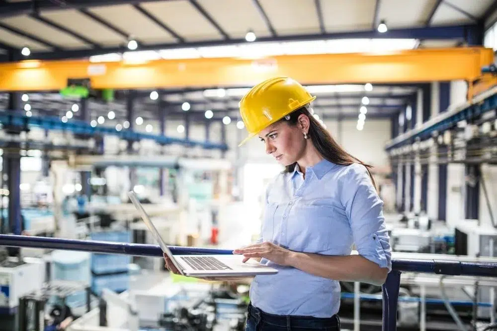 A person in a yellow hard hat uses a laptop while overlooking a busy industrial facility with machinery in the background.