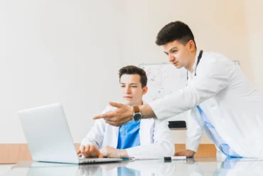 Two healthcare professionals in white lab coats discuss information while observing a laptop in a bright, modern office setting.