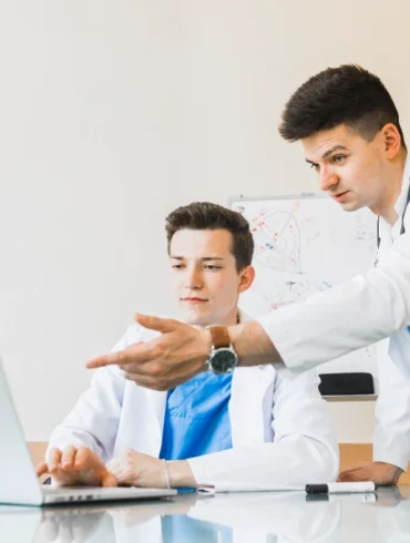 Two healthcare professionals in white lab coats discuss information while observing a laptop in a bright, modern office setting.