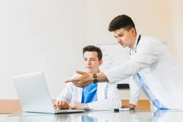 Two healthcare professionals in white lab coats discuss information while observing a laptop in a bright, modern office setting.