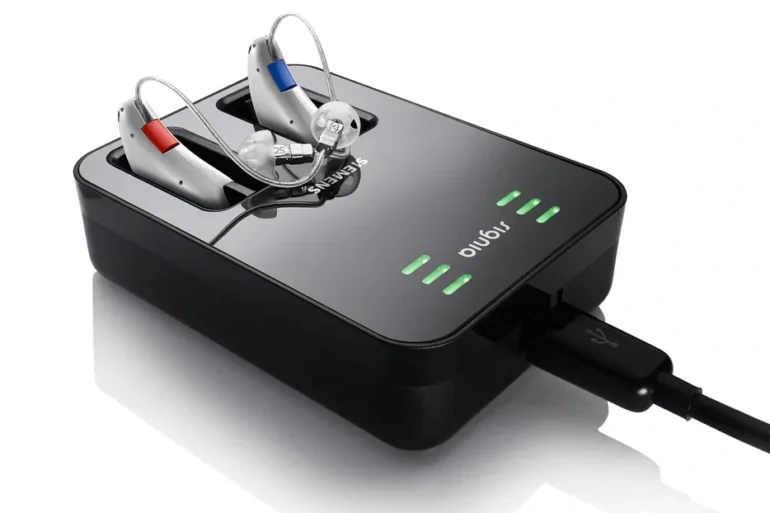 Charging dock with two hearing aids, one with a red and the other with a blue indicator, connected via USB cable.