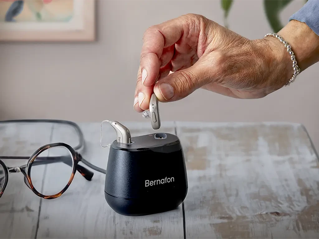 A hand places a hearing aid into a charging dock, with eyeglasses nearby on a wooden surface. The dock is labeled "Bernafon."