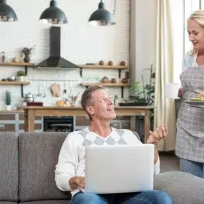 A man with a laptop sits on a couch, looking up smiling at a woman in an apron holding a plate and mug. A cozy kitchen is visible in the background.
