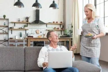 A man with a laptop sits on a couch, looking up smiling at a woman in an apron holding a plate and mug. A cozy kitchen is visible in the background.