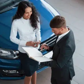 A business meeting in a car dealership, with a salesperson discussing details with a client near a blue vehicle.