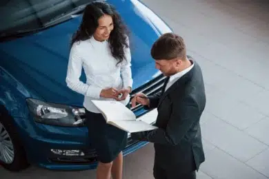 A business meeting in a car dealership, with a salesperson discussing details with a client near a blue vehicle.