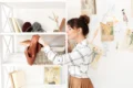A woman in a white plaid shirt places a brown fabric in a woven basket on a white bookshelf. Yarn, sketches, and fashion illustrations adorn the wall. The setting is bright and creative.