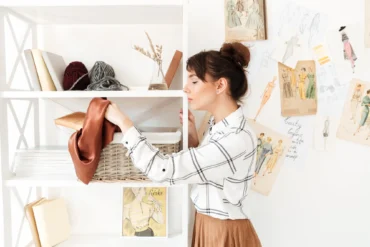 A woman in a white plaid shirt places a brown fabric in a woven basket on a white bookshelf. Yarn, sketches, and fashion illustrations adorn the wall. The setting is bright and creative.