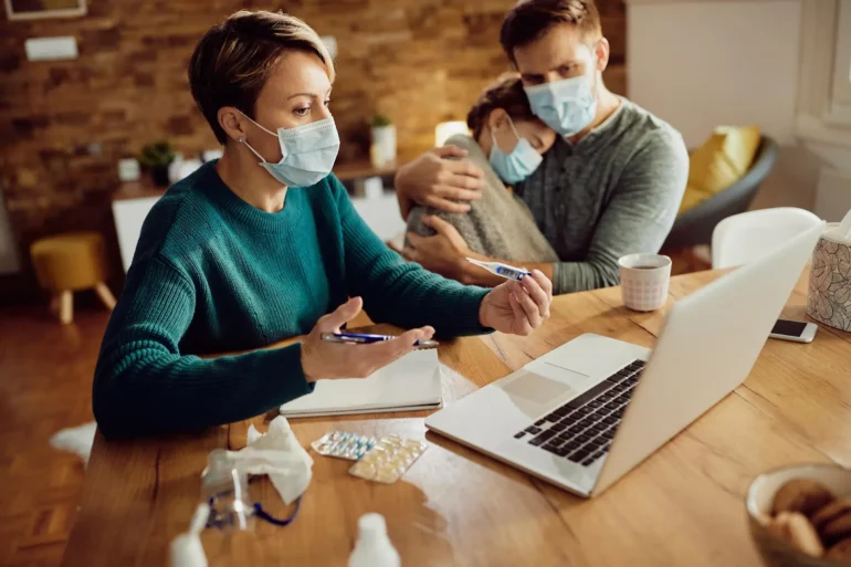 A woman in a teal sweater wearing a mask holds a pen, gesturing toward a laptop on a table with medicine. Behind, a masked man comforts a child.