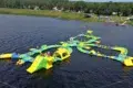 A large inflatable water park floats on a lake, featuring slides, obstacles, and climbing structures in green and yellow. People play under a clear sky.