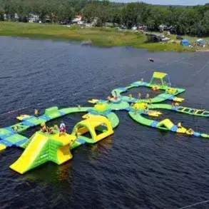 A large inflatable water park floats on a lake, featuring slides, obstacles, and climbing structures in green and yellow. People play under a clear sky.