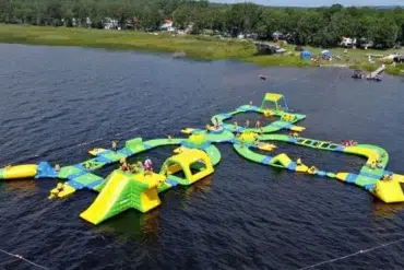 A large inflatable water park floats on a lake, featuring slides, obstacles, and climbing structures in green and yellow. People play under a clear sky.