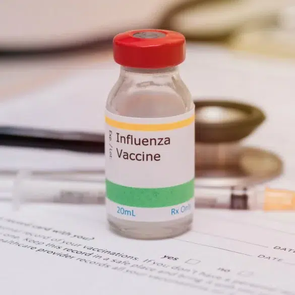 A vial labeled "Influenza Vaccine" with a red cap is placed on a medical form. A syringe and stethoscope lie nearby, suggesting a medical setting.