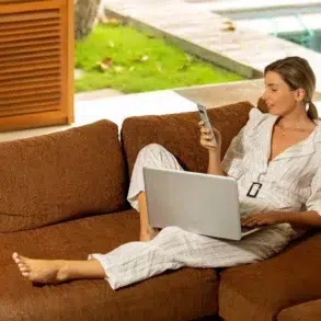 Woman in a relaxed setting, reclining on a brown sofa, using a smartphone in one hand and a laptop on her lap. She's wearing a striped outfit, with a view of a garden and a swimming pool in the background. The ambiance is calm and focused.